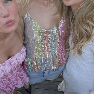 Glittrig paljettlinne i regnbågsfärger - Säljer ett glittrigt linne med paljetter i regnbågsfärger. Linnet har smala axelband och en lös passform, perfekt för en festlig kväll. Färgerna skiftar i rosa, grönt, guld och silver vilket ger en iögonfallande look.