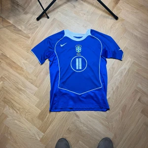 Blå Brasilien fotbollströja med nummer 11 och Ronaldinho  - Säljer en blå Brasilien fotbollströja från Nike med nummer 11 och texten 'Ronaldinho' på ryggen. Tröjan har korta ärmar och vita detaljer. Perfekt för fotbollsfans!