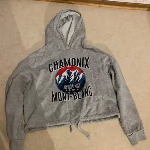 Säljer en grå hoodie med tryck av Chamonix Mont-Blanc och bergsmotiv. Hoodien har en croppad design med dragsko i nederkanten och långa ärmar. Perfekt för en avslappnad stil.