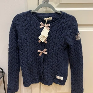 Mörkblå stickad kofta med rosetter - Charmig mörkblå stickad kofta med dekorativa rosetter framtill. Koftan har ett flätmönster och en broderad detalj på ärmen. Perfekt för kyliga dagar.