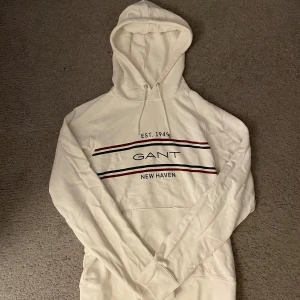 Vit hoodie från GANT - Säljer en stilren vit hoodie från GANT med tryck i rött och blått. Hoodien har en klassisk design med dragsko i huvan och en stor ficka framtill. Storlek L men sitter som M. En av metallbitarna till ett av snörena e borta, se sista bilden. Annars inga defekter. Hör av er vid funderingar:)