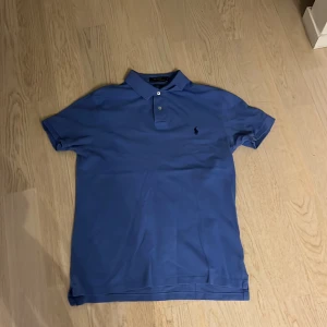 Blå pikétröja från Ralph Lauren - Säljer en stilren blå pikétröja från Ralph Lauren i slim fit. Tröjan har korta ärmar och en klassisk krage med knappar. Den ikoniska loggan är broderad på bröstet. Perfekt för sommaren och våren.