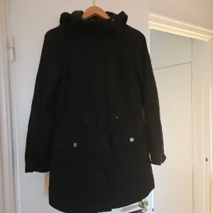 Parkas nyskick  - Nyskick Svart parkas med huva. Jackan har dragsko i midjan och fickor med tryckknappar och dragkedja. Ärmarna har justerbara tryckknappar.