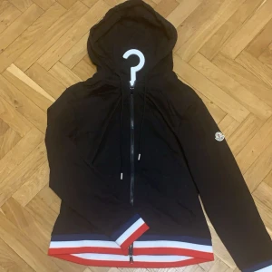 Svart hoodie från Moncler - Snygg svart hoodie från Moncler med dragkedja och huva. Den har en broderad Moncler-logga på ärmen och ribbade muddar i rött, vitt och blått vid nederkanten. Perfekt för en stilren look.