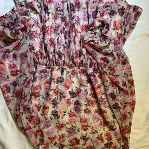 Blommig klänning med volangdetaljer - Charmig blommig klänning i rosa och lila toner med volangdetaljer. Perfekt för sommarens alla tillfällen. Den har en luftig passform och är tillverkad i ett lätt material som ger en skön känsla.