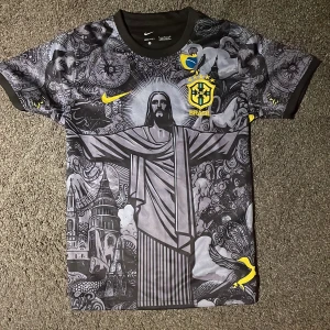 Svart Brasilien-jesus fotbollströja från Nike - Snygg svart fotbollströja från Nike. Tröjan har ett unikt mönster med Kristusstatyn och andra detaljer i grått och gult. Den är kortärmad och tillverkad i Dri-FIT-material för optimal komfort.