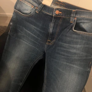 Blå jeans från Nudie Jeans - Snygga blå jeans från Nudie Jeans med klassisk femficksdesign och orange sömmar. Jeansen har en dragkedja och knapp framtill samt en diskret logga på bakfickan. Perfekta för en avslappnad stil.
