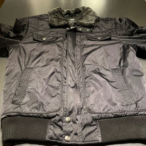 Svart jacka från Giorgio Armani - Säljer en stilren svart jacka från Giorgio Armani med fuskpäls på kragen. Jackan har dragkedja och knappar framtill samt ribbade muddar. Den är tillverkad i syntetmaterial och har en cool detalj med metallspännen på sidorna. En av dem är trasiga 