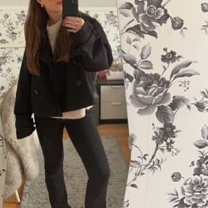 Svart kort trenchcoat - Snygg svart kappa med dubbelknäppning och långa ärmar. Jackan har en croppad design och är perfekt för höst och vår. Den har en stilren look som passar till många olika outfits.