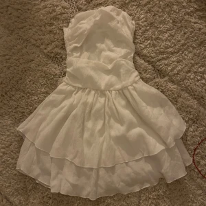 Vit axelbandslös klänning - Söt vit axelbandslös klänning med volangdetaljer. Perfekt för sommarens festligheter. Den har en kort längd och en luftig passform 