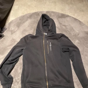 Svart hoodie med dragkedja  - Säljer en svart hoodie med dragkedja och en bröstficka. Perfekt för kyliga dagar med sin bekväma passform och huva. En stilren design som passar till det mesta.