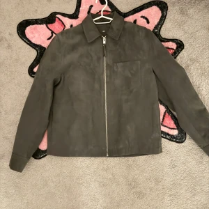 Grå mockajacka från H&M - Snygg grå mockajacka från H&M med dragkedja framtill och en bröstficka. Jackan har en klassisk krage och långa ärmar med knappdetaljer vid ärmsluten. Perfekt för en stilren look.