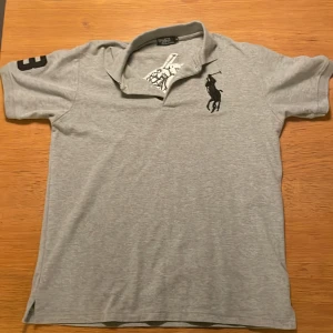 Grå pikétröja från Polo - Säljer en klassisk grå pikétröja från Polo i 100% bomull. Tröjan har korta ärmar och en broderad logga på bröstet samt en stor siffra på ärmen. Inga fläckar den är som ny. Pris kan diskuteras.
