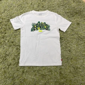 Vit t-shirt från Levi's med tryck - Säljer en vit t-shirt från Levi's med ett coolt tryck i grönt och gult. T-shirten har korta ärmar och är perfekt för en avslappnad stil. Den är gjord i mjuk bomull och har en normal passform.