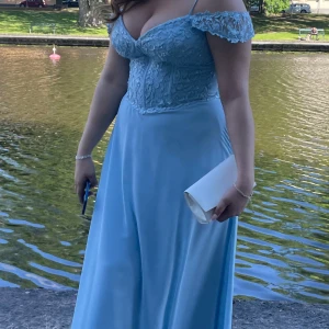 Ljusblå långklänning med spetsdetaljer - Elegant ljusblå långklänning med offshoulder-design och spetsdetaljer på överdelen. Klänningen har en vacker fall och smala axelband, perfekt för en bal eller festlig tillställning. Kombinera med en stilren clutch för en komplett look. 