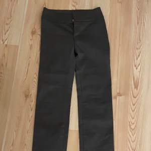 Svarta byxor från Monki - Snygga svarta byxor från Monki i storlek 36. Byxorna har en rak passform och är tillverkade i ett syntetmaterial. De har en stilren design med knappar i midjan och en diskret slits vid benslutet.