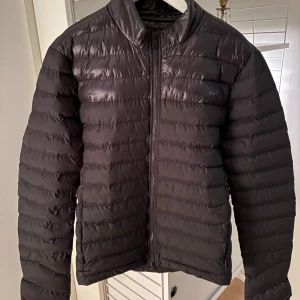 Svart dunjacka från J.Lindeberg - Snygg svart dunjacka från J.Lindeberg med dragkedja framtill. Jackan har en quiltad design och är perfekt för kyligare dagar. Den har en stilren look och passar bra till olika outfits.