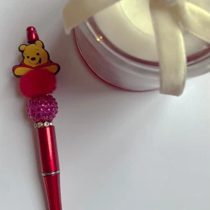 Röd penna med Nalle Puh-design❤️ - En röd penna dekorerad med en Nalle Puh-figur på toppen. Pennan har en glittrig sektion med rosa stenar och en fluffig röd boll. Perfekt för att lägga till lite lekfullhet till skrivbordet.❤️