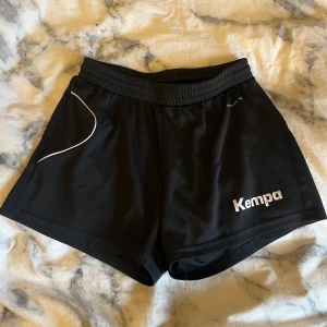 Svarta shorts från Kempa - Snygga svarta shorts från Kempa med elastisk midja, snören för att få ännu tajtare och vit logga på framsidan och sidan. Perfekta för sport och träning med en bekväm passform.