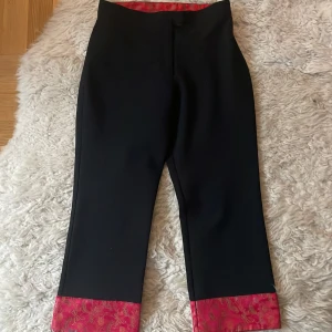 Svarta capri byxor med röd detalj st.164 - Snygga svarta byxor med en unik röd och blommig detalj vid bensluten. Byxorna har en rak passform och är perfekta för att ge en stilren look med en twist. Den röda färgen ger en fin kontrast och gör dem speciella.