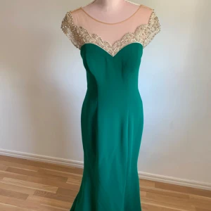 Grön långklänning med pärlor - Elegant grön långklänning med en vacker pärlbroderad överdel och transparent mesh. Klänningen har en figurnära passform och en dragkedja i ryggen. Perfekt för festliga tillfällen.