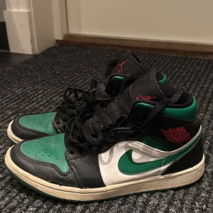 Nike Air Jordans i svart och grön - Snygga Nike Air Jordans i svart och grön med vita detaljer. Skorna har en klassisk hög design med snörning och den ikoniska Air Jordan-loggan i rött på sidan. Perfekta för dig som vill ha en stilren och sportig look.