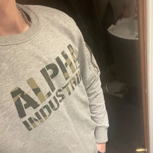 Grå sweatshirt från Alpha Industries - Säljer en grå sweatshirt från Alpha Industries med camouflagemönstrad logga på bröstet. Tröjan har långa ärmar och ribbade muddar vid ärmslut och nederkant. Perfekt för en avslappnad stil. Skriv gärna vid minsta fundering!