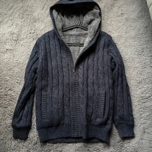 Mörkblå stickad hoodie med dragkedja - Säljer en mysig mörkblå stickad hoodie med dragkedja och huva. Tröjan har ett kabelstickat mönster och är perfekt för kyliga dagar. Den är helt oanvänd.