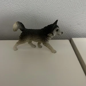 Schleich hund - En liten figur av en hund med detaljerad pälsstruktur. Färgerna är främst svart och vit med grå nyanser. Figuren står i en naturlig pose med svansen uppåt.