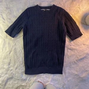 Mörkblå stickad tröja - Säljer en stilren mörkblå stickad tröja med korta ärmar. Perfekt för en avslappnad look. Tröjan har ett diskret mönster och ribbade kanter vid ärmslut och nederkant.