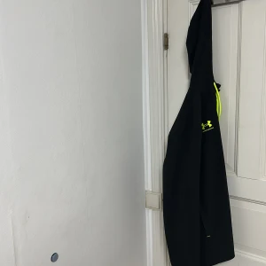 Svart träningsjacka från Under Armour - Snygg svart träningsjacka från Under Armour med huva och gula detaljer. Perfekt för sportiga aktiviteter. Jackan har långa ärmar och en bekväm passform.