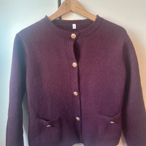 Vinröd kofta med knappar - Säljer en mysig lila kofta med dekorativa knappar framtill. Koftan har långa ärmar och två små fickor. Perfekt för kyliga dagar.