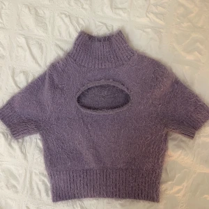 Lila stickad tröja från Collusion - Säljer en lila stickad tröja från Collusion med en unik cut-out detalj framtill. Tröjan har en hög krage och korta ärmar, perfekt för lager-på-lager eller som statement-plagg. Supermjuk och bekväm!