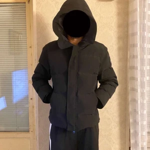 Svart pufferjacka med huva - Snygg svart pufferjacka med huva och broderad detalj på ärmen. Jackan har långa ärmar och ribbade muddar för extra komfort. Perfekt för kyliga dagar.