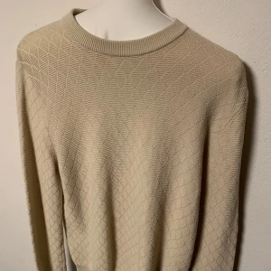 Beige stickad tröja från These Glory Days - Säljer en stilren beige stickad tröja med ett diskret rutmönster. Tröjan har långa ärmar och ribbade muddar vid ärmslut och nederkant. Perfekt för kyligare dagar. Storlek M