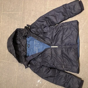 Mörkblå dunjacka från GANT - Snygg mörkblå dunjacka från GANT med huva och dragkedja. Perfekt för kyliga dagar. Jackan har en stilren design med broderad logga på bröstet och är tillverkad i ett slitstarkt material. Storleken är 146-152 och för barn mellan 11-12 år.