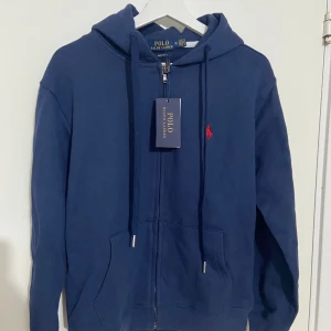 Blå hoodie zip från Ralph Lauren - Säljer en snygg blå hoodie från Ralph Lauren med dragkedja och det klassiska röda logotypen på bröstet. Perfekt för en avslappnad stil med justerbar huva och fickor framtill. Passar perfekt till både jeans och joggers.