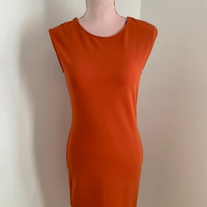 Orange klänning med spetsdetalj - Snygg orange klänning med en unik svart spetsdetalj på ryggen. Den är ärmlös och har en figurnära passform som framhäver silhuetten. Perfekt för en kväll ute eller en speciell tillställning.