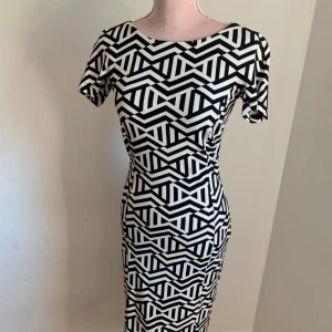 Svartvit mönstrad klänning med spetsdetalj - Snygg svartvit klänning med geometriskt mönster och korta ärmar. Klänningen har en unik spetsdetalj på ryggen som ger en elegant touch. Perfekt för att sticka ut med sin stilrena design.