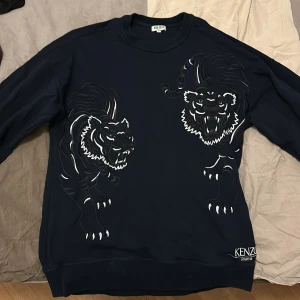 Marinblå sweatshirt från Kenzo med tigerdesign - Säljer en svart sweatshirt från Kenzo med en cool tigerdesign på framsidan. Tröjan har långa ärmar och en rund halsringning. Perfekt för dig som vill ha en stilren men ändå iögonfallande look. 🐯 Stor i storleken
