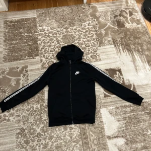 Svart huvtröja från Nike - Säljer en svart huvtröja från Nike med dragkedja och huva. Tröjan har långa ärmar med vita Nike-loggor längs sidorna. Perfekt för en sportig look eller avslappnad stil.