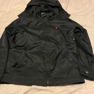  Ralph Lauren jacka - Säljer en stilren svart jacka från Ralph Lauren med huva och dragkedja. Jackan har långa ärmar och två fickor med knappar framtill. Den ikoniska loggan syns i rött på bröstet. Perfekt för en casual look.