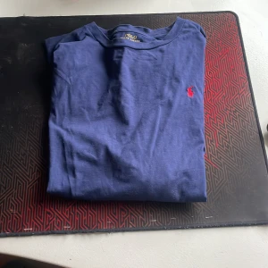 Blå t-shirt från Ralph Lauren - Säljer en klassisk blå t-shirt från Ralph Lauren med det ikoniska röda broderade logotypen på bröstet. Perfekt för en stilren och avslappnad look.