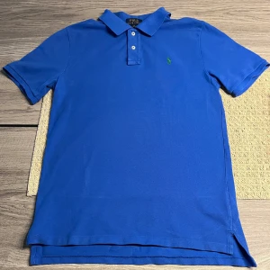 Blå pikétröja från Ralph Lauren - Säljer en klassisk blå pikétröja från Ralph Lauren med en broderad logotyp på bröstet. Tröjan har korta ärmar och en traditionell krage med knappar. Perfekt inför våren/sommaren.