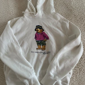 Vit hoodie med Polo Bear från Ralph Lauren - Säljer en vit hoodie från Ralph Lauren med det ikoniska Polo Bear-motivet. Björnen har en rosa tröja och mönstrade shorts. Perfekt för en avslappnad stil. 🐻nästan inte andvänd alls 