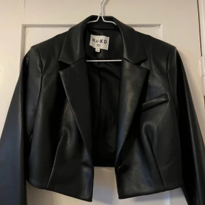Svart skinnjacka från NA-KD - Snygg svart skinnjacka från NA-KD i croppad modell. Jackan har en knapp framtill och klassisk krage. Perfekt för att ge din outfit en edgy touch.