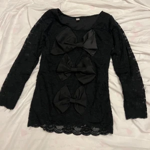 Svart spetsblus med rosetter storlek S oanvänd  - Snygg svart spetsblus med tre stora rosetter framtill. Blusen har långa ärmar och en elegant design som passar perfekt för speciella tillfällen. Den är tillverkad i ett vackert spetsmaterial som ger en lyxig känsla.