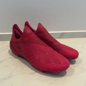 Röda fotbollsskor från Adidas - Säljer ett par röda fotbollsskor från Adidas i mycket bra skick. Skorna har en strömlinjeformad design utan snörning för en tight passform. Perfekta för fotbollsspelare som vill ha stil och funktion på planen.