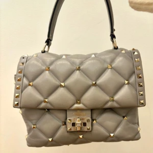 Valentino candystud väska - Jätte snygg ljusgrå Valentino Garavani väska, bra skick men lite sliten som man ser på bilderna och två nitar har ramlat av. Lite sminkfläckar inuti väskan se bilderna. Finns ett axelband till som medföljer. Nypris var ca 23000kr. Kvitto finns!❤️