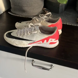 Nike fotbollsskor  - Säljer ett par Nike fotbollsskor i vitt med röda och svarta detaljer. Skorna har snörning och är i bra skick, perfekta för fotbollsträning eller match. De har en snygg design med 'AIR' tryckt på sidan och en svart swoosh på framsidan. Passar dig som vill ha stil och funktion på planen!